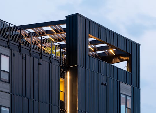A modern building with a dark facade and a large window with a light shining through.