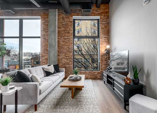 A living room with a grey couch, a wooden coffee table, and a large window with a view of a tree.at Raymond Block, Edmonton Alberta T6E 3R7