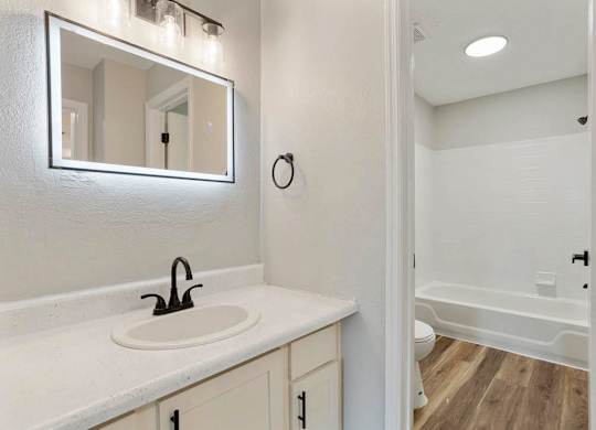 A white bathroom with a sink, mirror, and wooden floor at The Onyx Hoover Apartments, Alabama