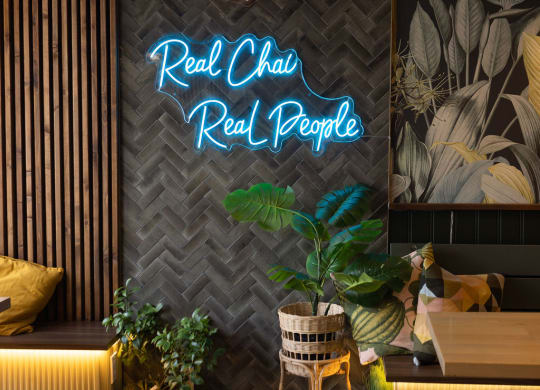 a neon sign that reads real chic real people on a wall with plants in front of it