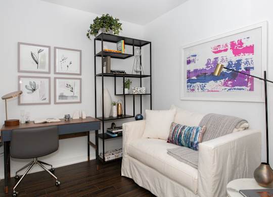 A living room with a white couch, a desk with a chair, and a bookshelf with a plant on top.