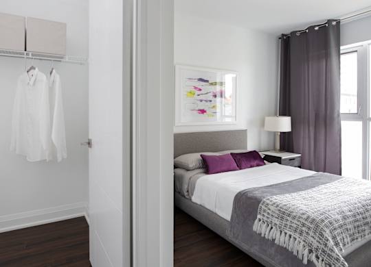 A bedroom with a bed, a nightstand, a lamp, a painting, a window with curtains, a hanging shirt, and a wall-mounted coat rack.