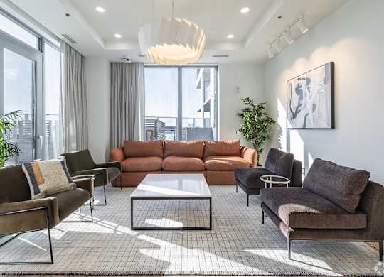 A living room with a brown sofa, a grey armchair, a brown chair, a glass coffee table, a potted plant, and a painting on the wall.