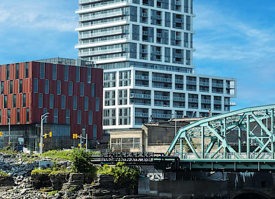 A modern glass building stands tall next to a red brick building with a bridge crossing a river in the foreground.