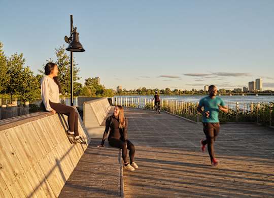 A woman in a white shirt and black pants is sitting on a wooden wall while another woman in a blue shirt and black pants is running on the boardwalk.