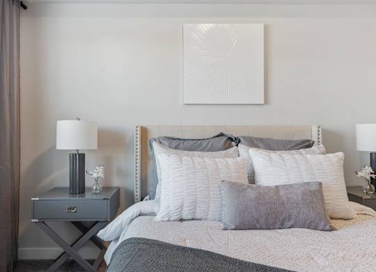 a bedroom with a bed and pillows and a nightstand with a lamp and at The Flats at Riverwoods, Provo, UT, 84604