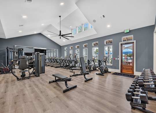 A gym with a variety of equipment including treadmills, weights, and a bench press. Large well lit room with a fan and tvs