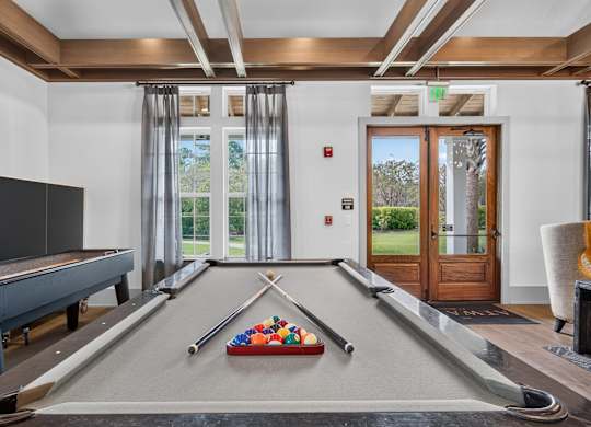 A pool table with a red cloth and a set of pool balls in the corner of a room with a door and a window, in the clubhouse next to the lounge