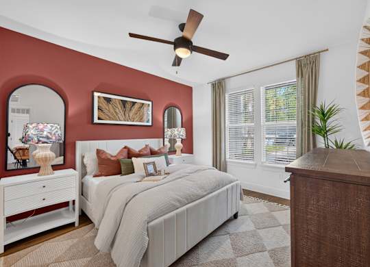 A bedroom with a large bed, a dresser, and a ceiling fan, the wall behind the bed is painted a soft red with a mirror above the bedside table