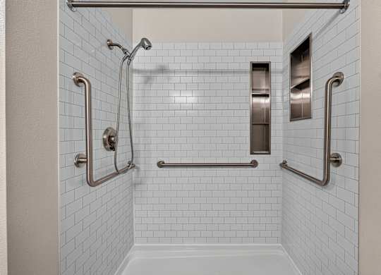 A white tiled shower with a glass door and a metal grab bar. Steel fixtures updated shower with  storage.