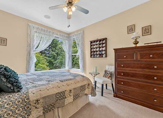 A bedroom with a large bed and a dresser. Blue bedding. Lace curtains over large windows. Ceiling fan with the light on. Artwork on the walls