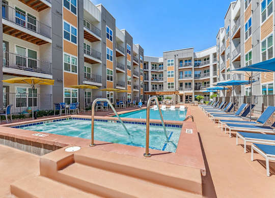 A large outdoor pool surrounded by lounge chairs and umbrellas in front of apartment buildings.