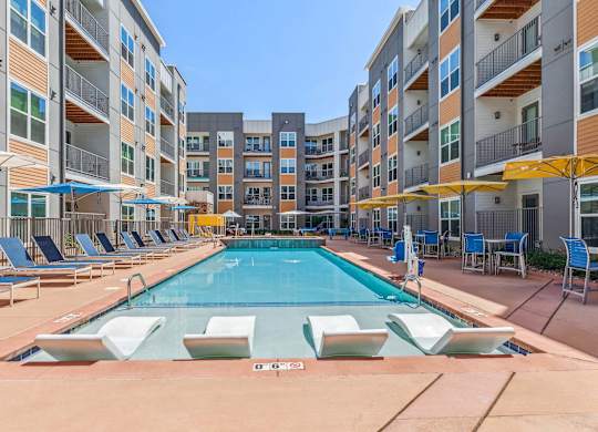A large swimming pool surrounded by lounge chairs and umbrellas in front of apartment buildings.