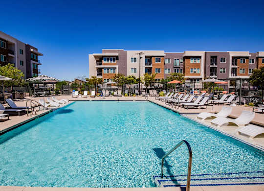 A large swimming pool surrounded by sun loungers and umbrellas in front of apartment buildings.