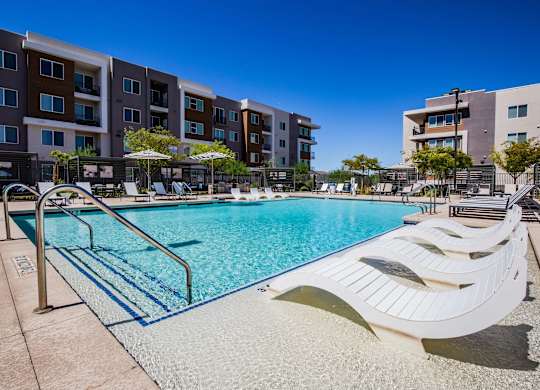 A swimming pool surrounded by lounge chairs and umbrellas in front of apartment buildings.