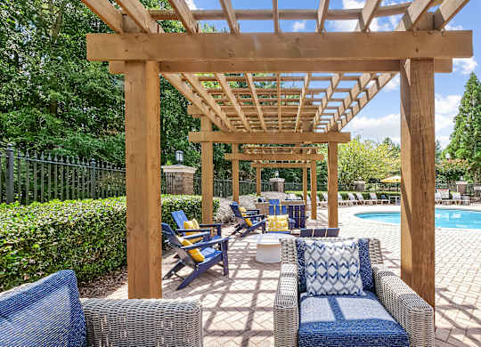 A wooden pergola over a poolside seating area with blue cushioned chairs.