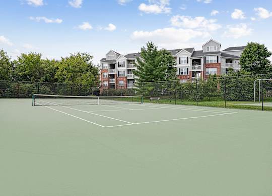 A tennis court with a green surface and white lines, surrounded by a fence and with apartment buildings in the background.
