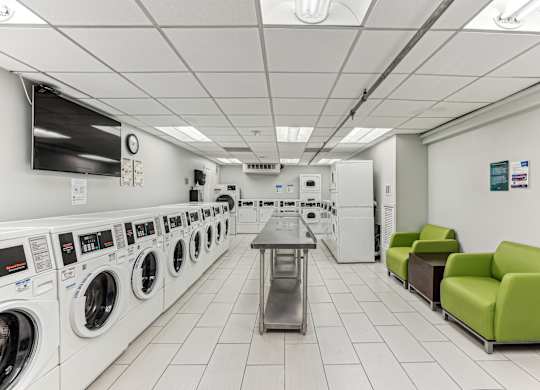 A laundromat with a row of washers and dryers on the left and a table and two chairs on the right.