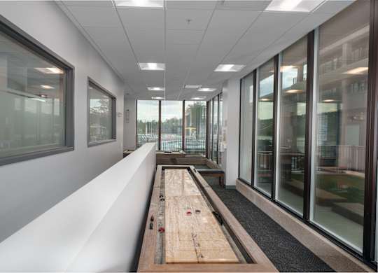 A long wooden table with a red object on it sits in a room with white walls and windows.