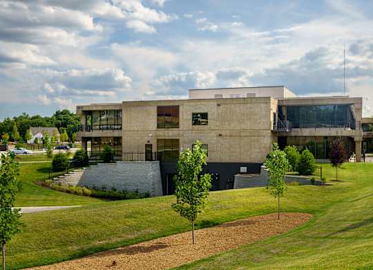A modern building with a flat roof and large windows is surrounded by a grassy area with trees.