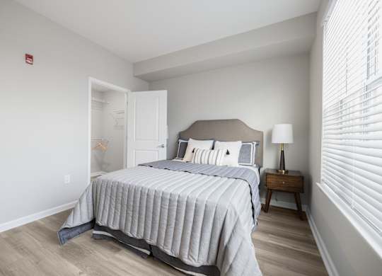 A bedroom with a bed, nightstand, and window with blinds at Harbor Pointe Apartments, New Jersey, 07002