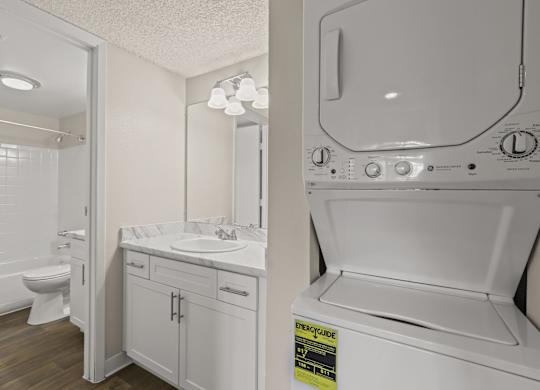 A white washing machine is in a laundry room with a sink and mirror at The Ranch at Bear Creek Apartments, Colorado, 80227