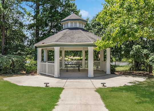 A gazebo with a white roof and pillars is surrounded by a grassy area and trees.