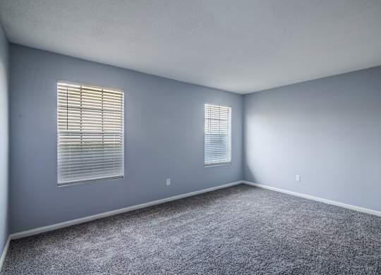 A room with grey carpet and two windows with blinds.