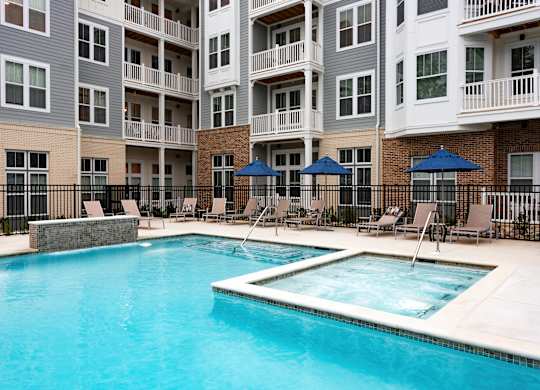 A swimming pool surrounded by chairs and umbrellas in front of apartment buildings.