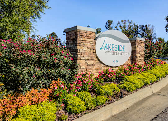 A sign for Lakeside Riverwatch stands in front of a flower bed.