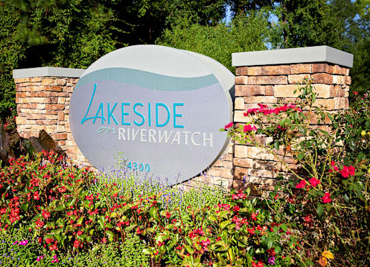 A sign that says Lakeside Riverwatch in front of some flowers.