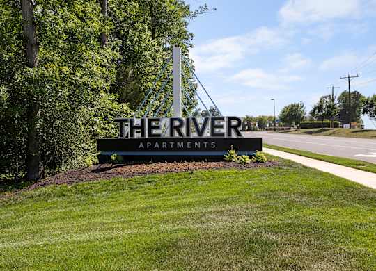 A sign for The River Apartments is displayed in front of a tree.