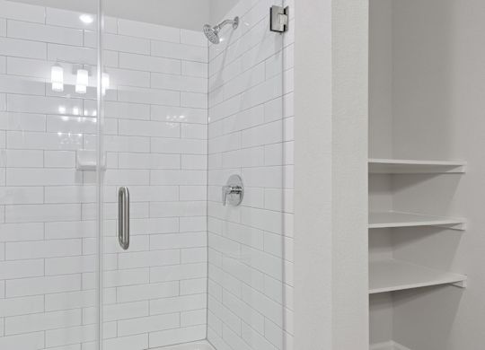 A white tiled shower with a glass door and a white shelf.