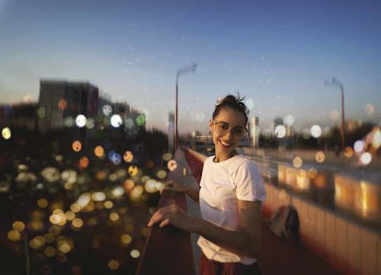 A woman in a white shirt and red skirt stands on a bridge with a cityscape in the background.