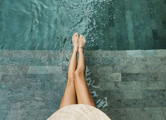 A person's legs are crossed at the ankles, wearing a straw hat, and standing on a stone ledge by a body of water.