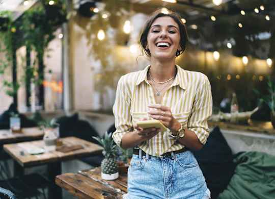 A woman in a striped shirt and jeans is laughing while holding a phone in a cozy, dimly lit room with hanging plants and tables.