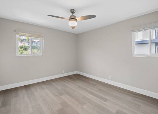A room with a ceiling fan and light, hardwood floors, and a window with a view of a building.
