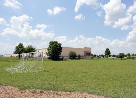 A soccer goal is in the foreground of a grassy field with a building in the background.