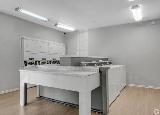 A white table with a grey countertop and a white cabinet with drawers is in a room with white walls and wooden flooring.