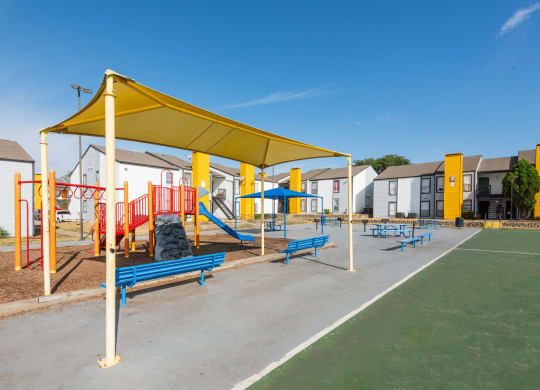a playground with benches and a yellow canopy in front of apartment buildings at Helios, Dallas, TX