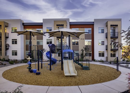 an outdoor playground with swings and umbrellas in front of apartments