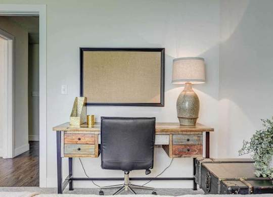 A black chair sits in front of a desk with a lamp and a picture frame on it.