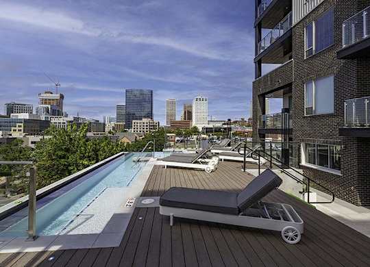 A rooftop pool with a lounge chair and a hot tub with a city skyline in the background.