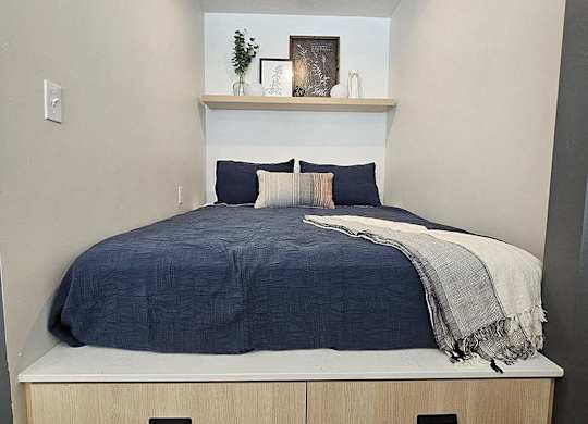 A bed with a blue comforter and pillows is in a room with a shelf above it.