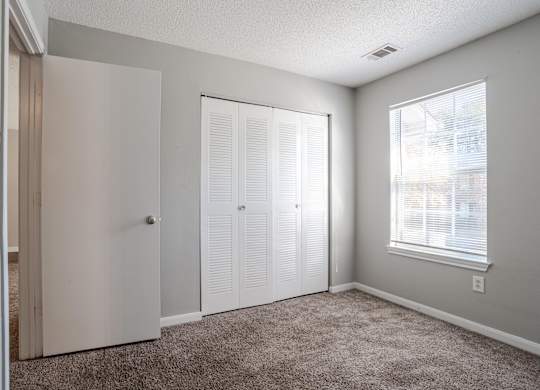 A room with a carpeted floor, a white door, a window with blinds, and a wall-mounted light fixture at Brookside Pointe Apartments, Travelers Rest, SC