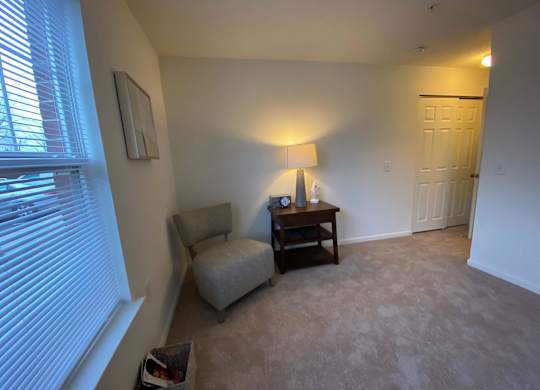 A living room with a chair, a table with a lamp and a clock on it, and a window with blinds.