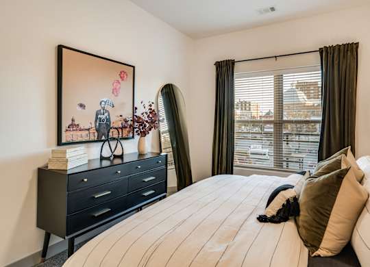 A bedroom with a large bed, a painting on the wall, and a window with blinds.at Converge KC Uptown, Kansas City, MO
