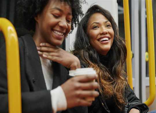 Two women are sitting on a bus and laughing while one of them holds a cup.