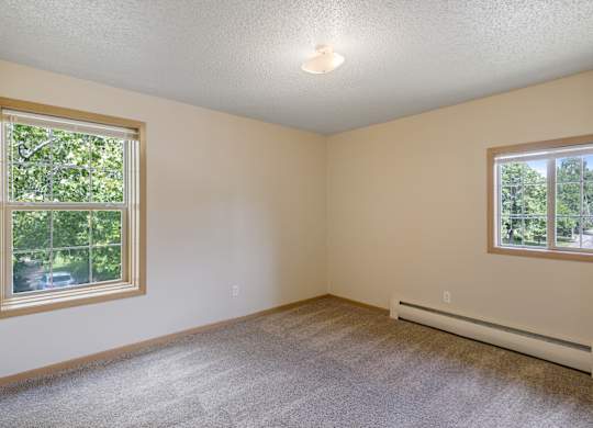 A room with a carpeted floor, a window with a view of trees, and a wall-mounted air conditioner.