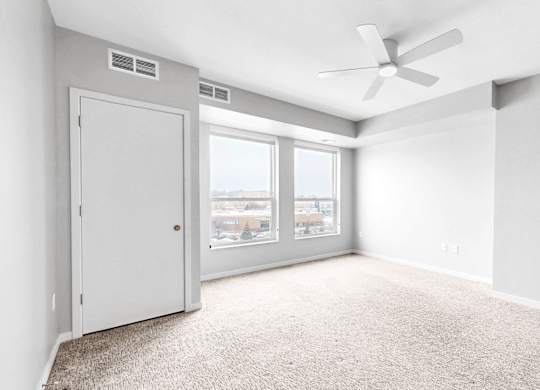A room with a carpet floor, a ceiling fan, and a window with a view of buildings outside.